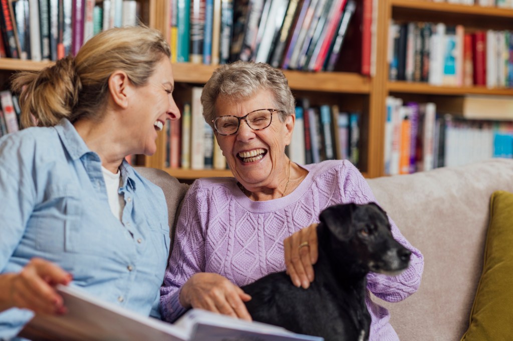 A mature woman sitting with her mother and pet dog on a sofa at home in Seghill, Northumberland. The senior woman is staying with her daughter for support and care as she has dementia and they are looking through a photo album together to aid her memory and strengthen their relationship.